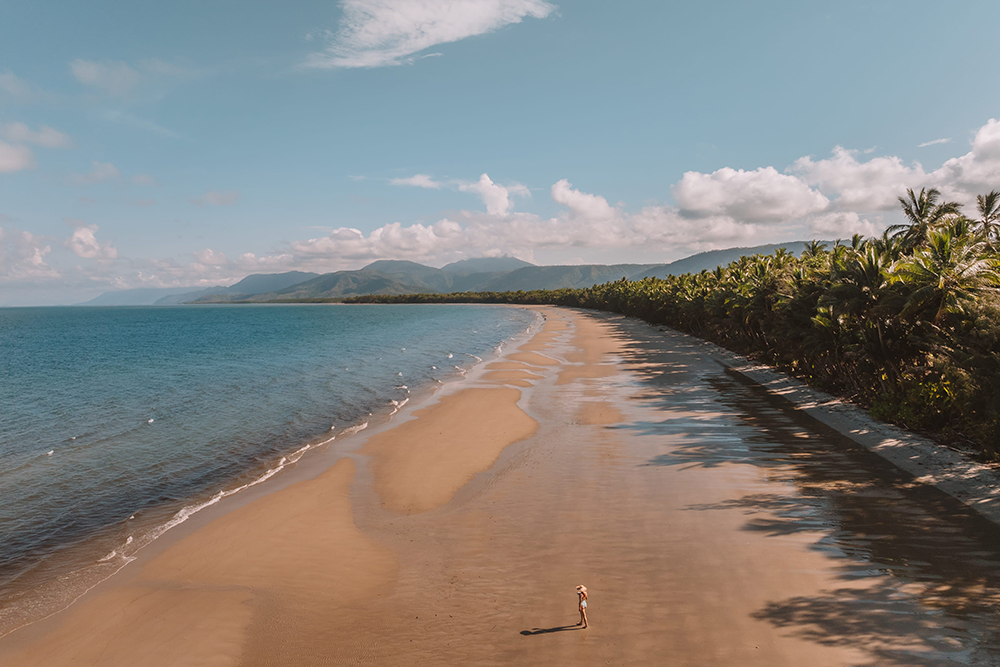 Lone figure on the wide sands of Four Mile Beach in Port Douglas, with palm trees and mountains in the distance
