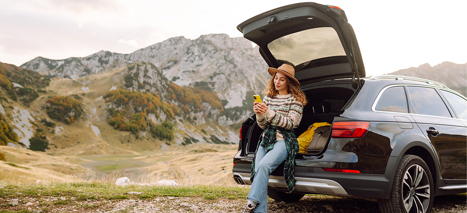 young woman sitting on the rear of a black car, smiling while using her phone in a mountainous area